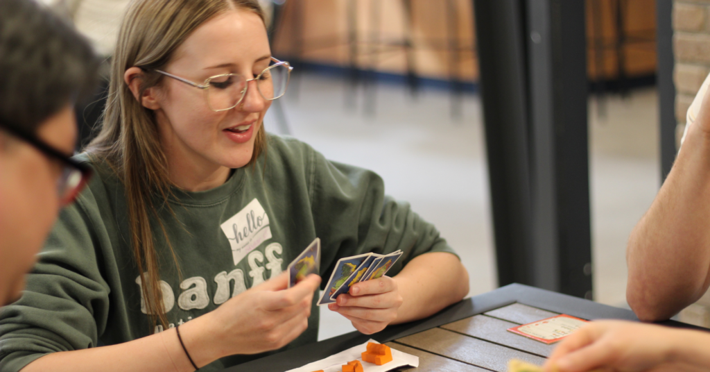 A woman smiling while holding Catan resource cards during a board game meetup in Saskatoon.